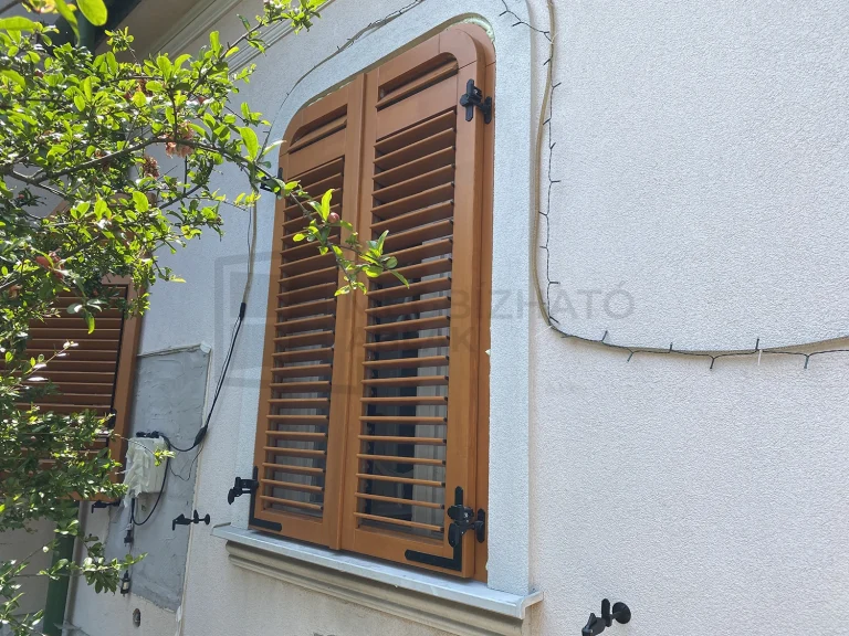 Világosbarna, kétszárnyú fa zsalugáter fekete vasalatokkal egy ablak előtt